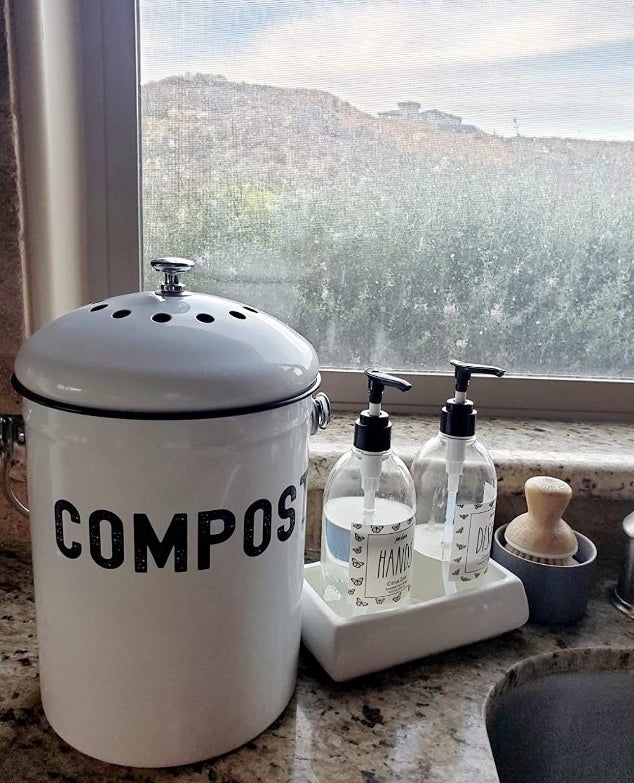 A white compost bin that says &quot;compost&quot; in black a in a reviewer&#x27;s home with hand and dish soap next to it