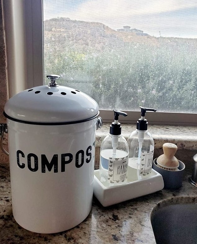 A white compost bin that says "compost" in black a in a reviewer's home with hand and dish soap next to it