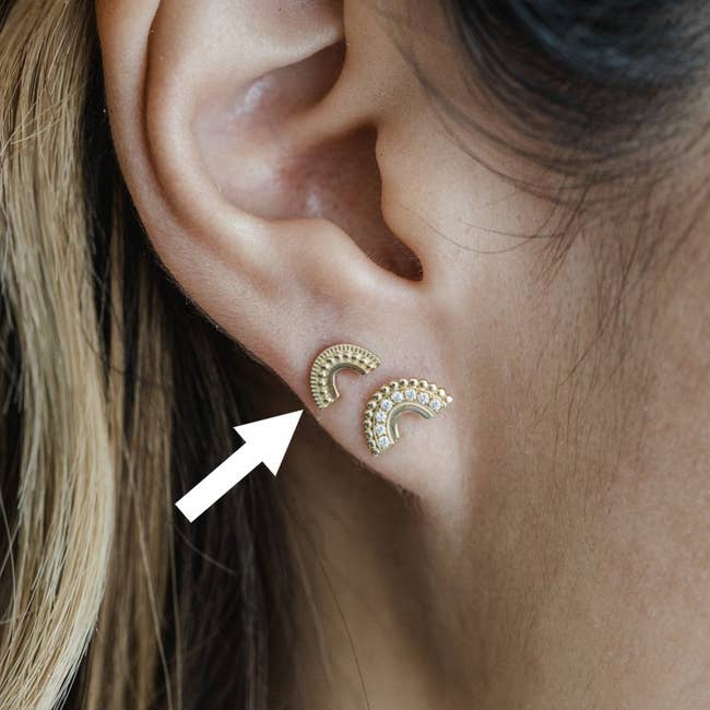 Image of model wearing two gold arc earrings with white arrow pointing to top earring