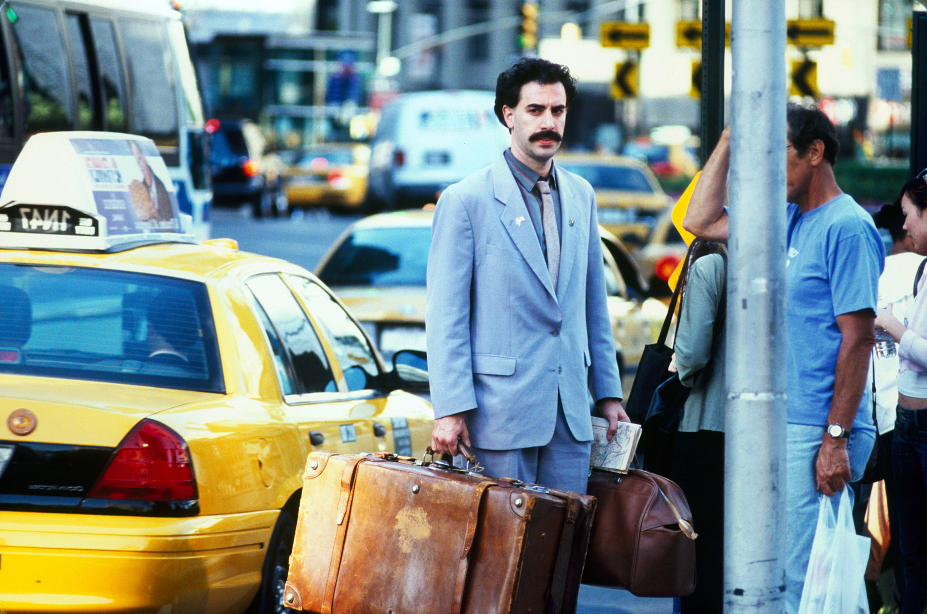 Sacha Baron Cohen as Borat standing in the middle of new york city with two suitcases