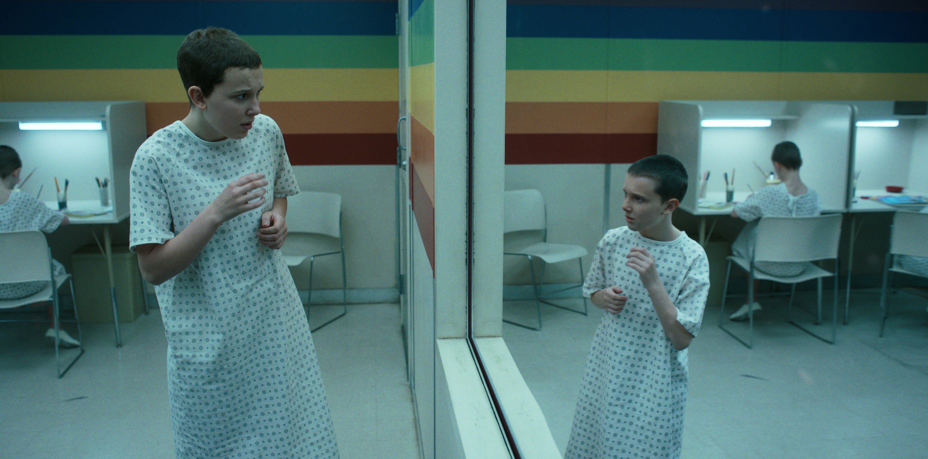 Eleven in a hospital gown looking in a mirror