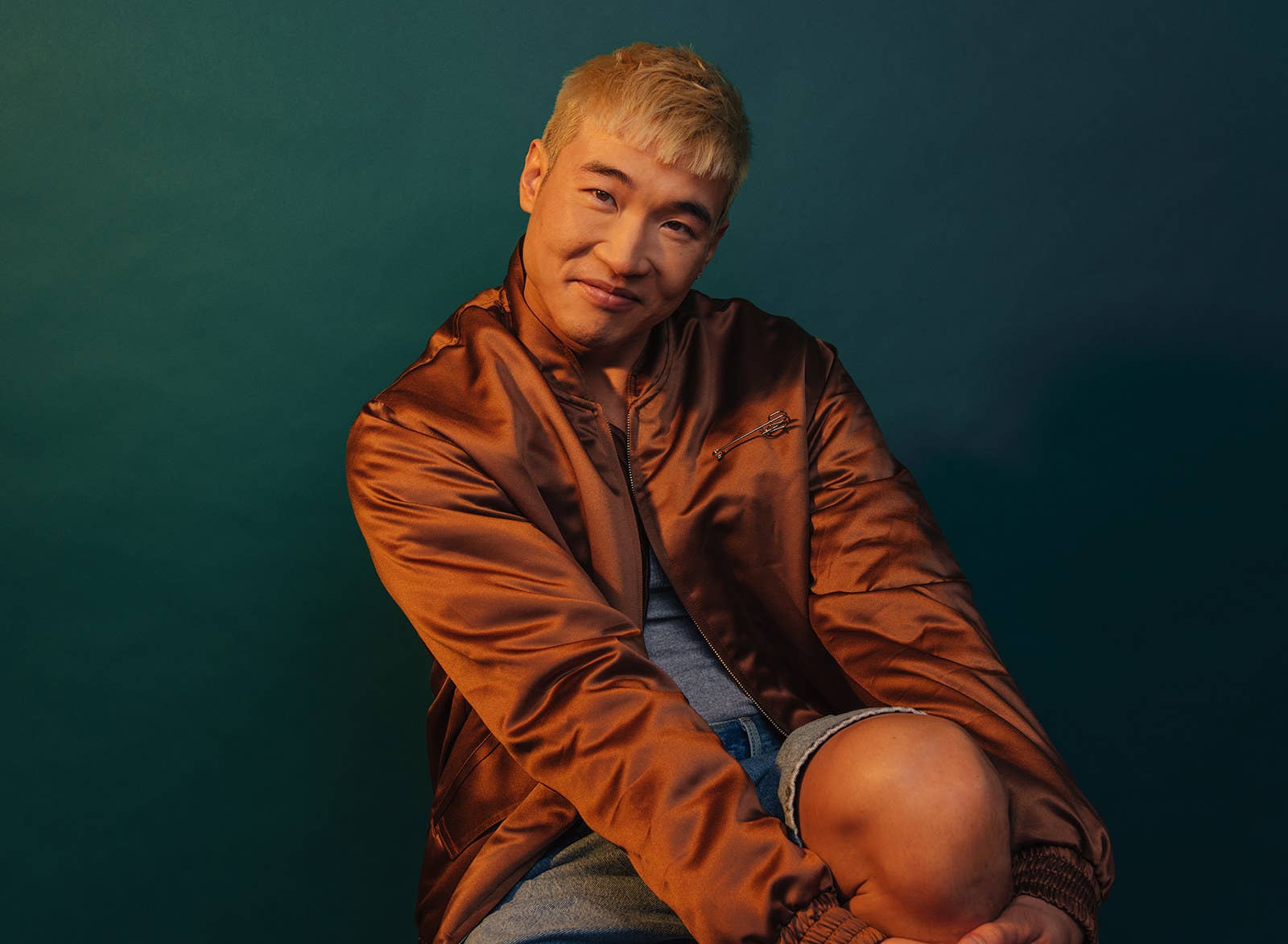 A man wearing a jacket and denim shorts sits in a photo studio for a portrait