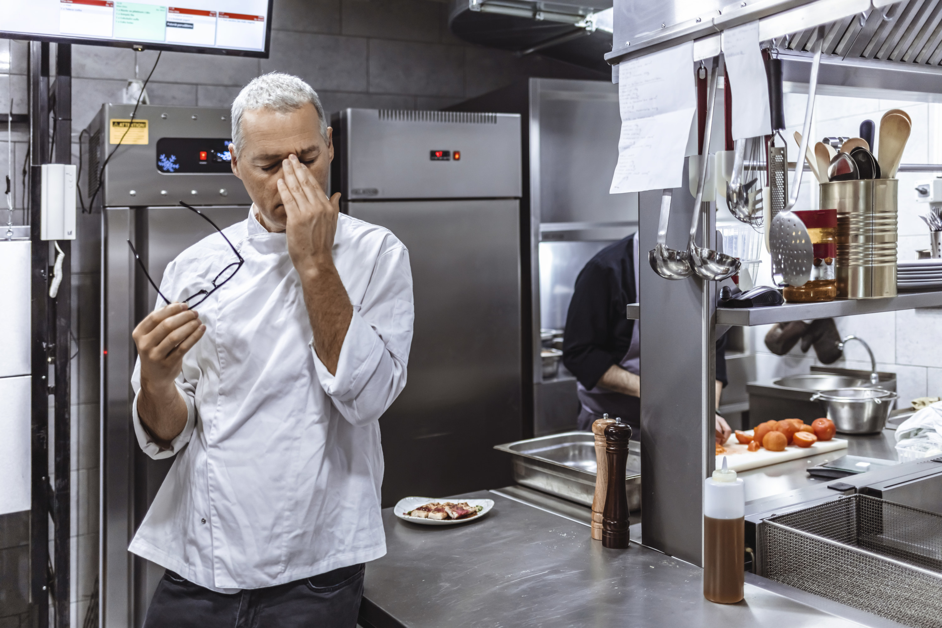 Person in a restaurant kitchen holding his eyeglasses and rubbing his eyes
