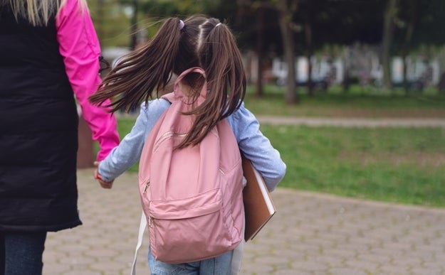 A young girl carries a backpack and her notebook to school with her mother by her side