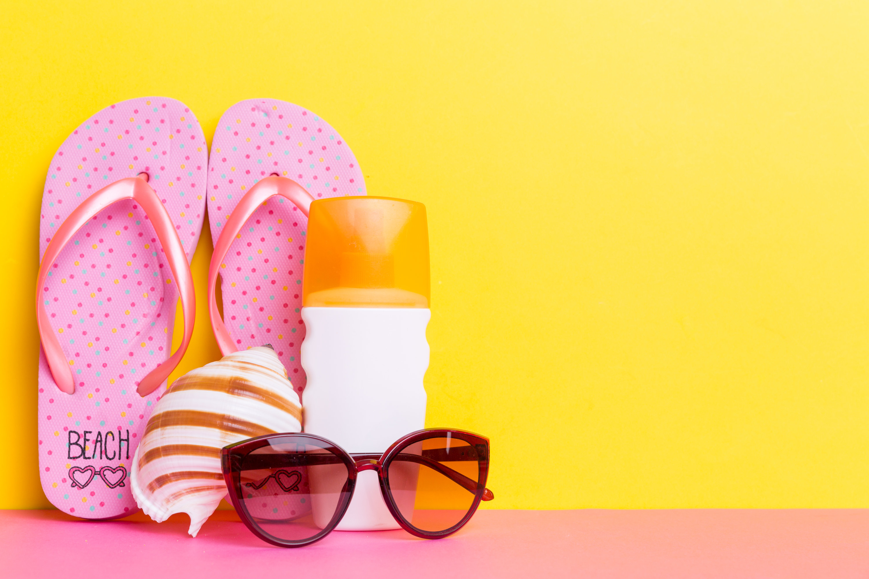 Beach accessories. Pink flip flops, sunglasses, sunscreen, and a sea shell propped against the wall