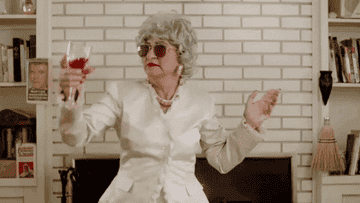 Old lady dancing and holding wine; She&#x27;s wearing a white suit and aviator sunglasses