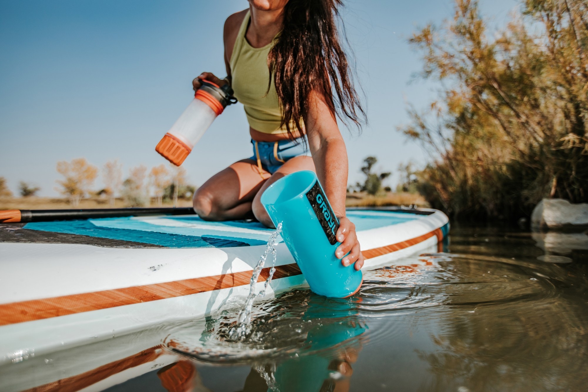 a model on a stand up paddle board getting water from a lake in the bottle