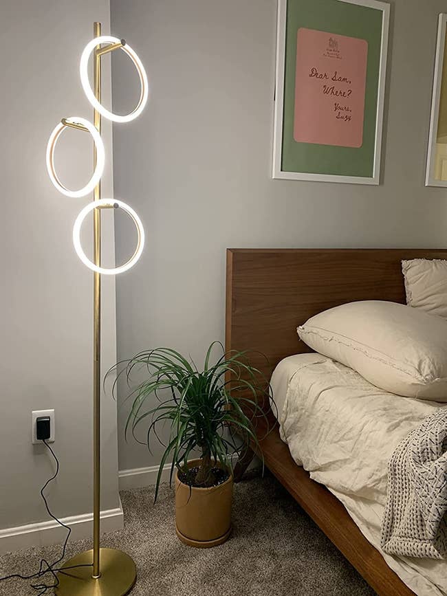 Modern floor lamp with four ring lights in a bedroom beside a plant and bed, with framed text artwork above the bed