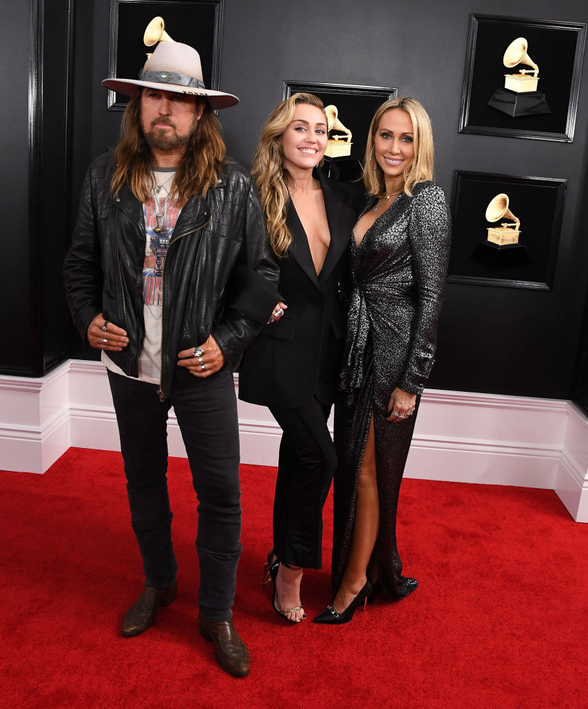 Billy Ray Cyrus wears dark jeans with a leather jacket, Miley Cyrus wears a dark suit with a v-neck, and Tish Cyrus wears a dark gown