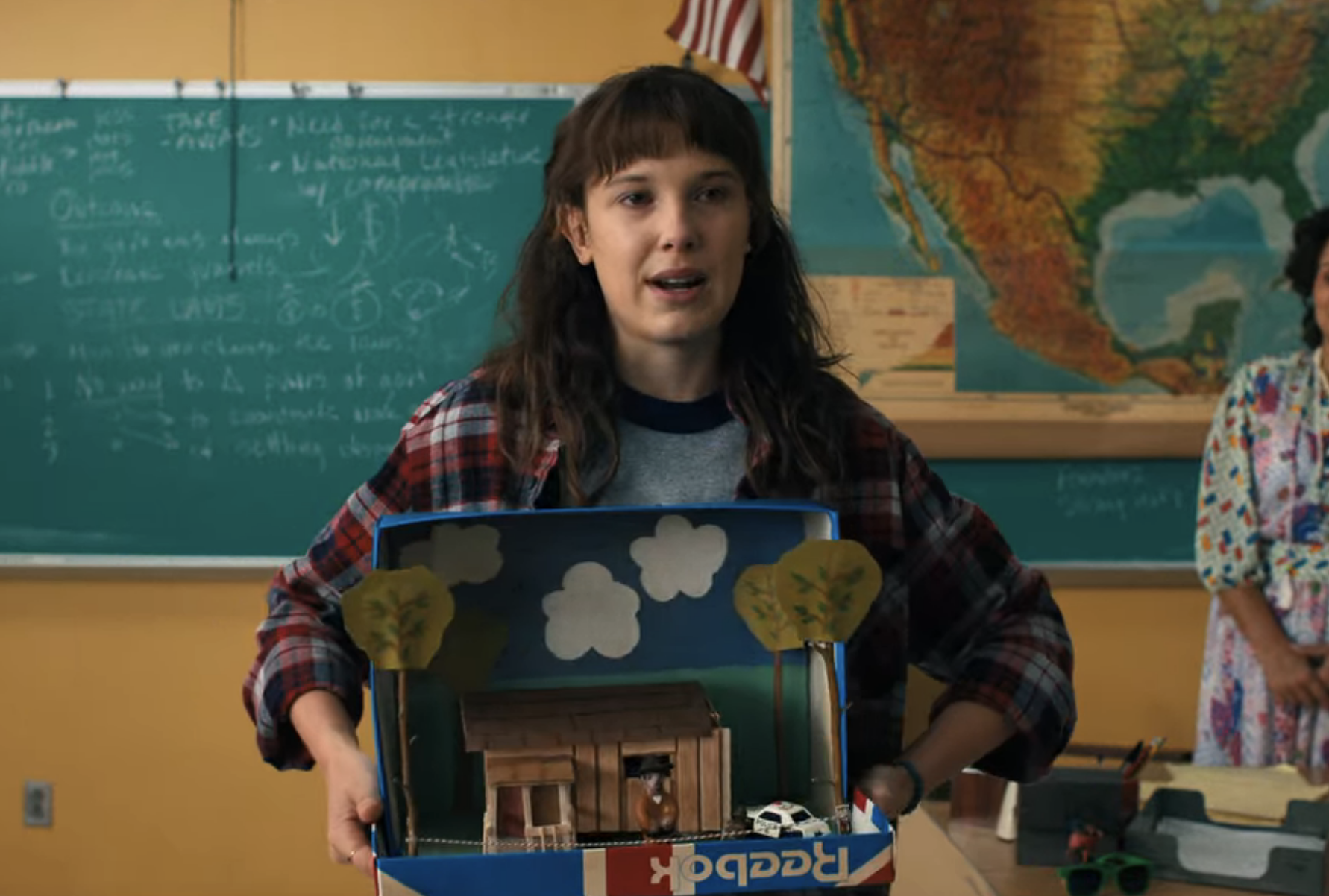 Eleven stands in front of the class holding a diorama