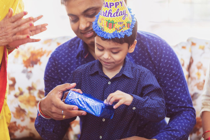 A son sitting on his father&#x27;s lap and unwrapping his birthday gift