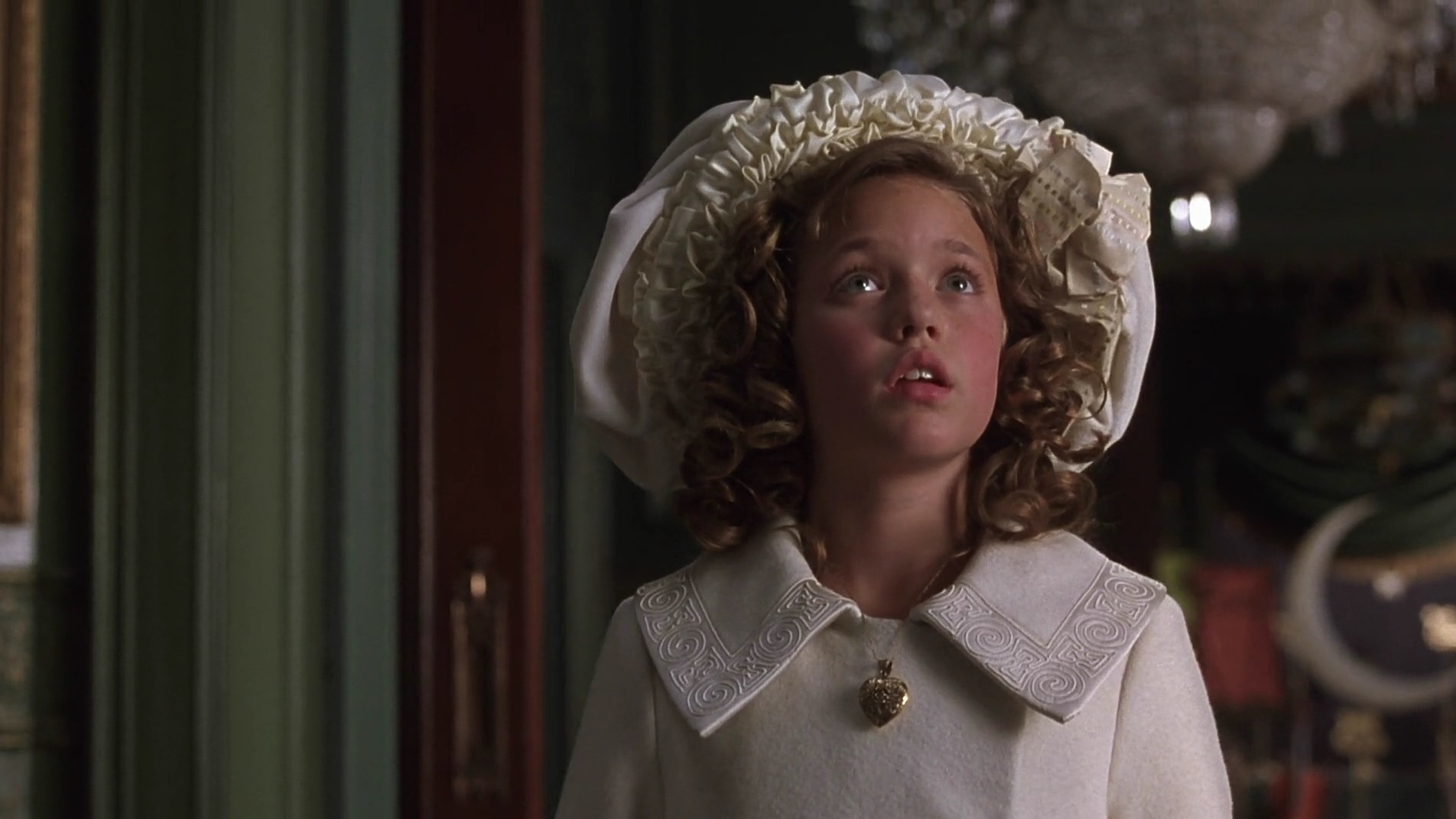 Sara wears an elaborate white hat, a white coat, and a gold locket, and stares upwards at something
