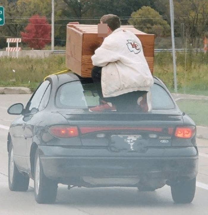 Person sitting on the trunk of a car to hold a dresser on top of it