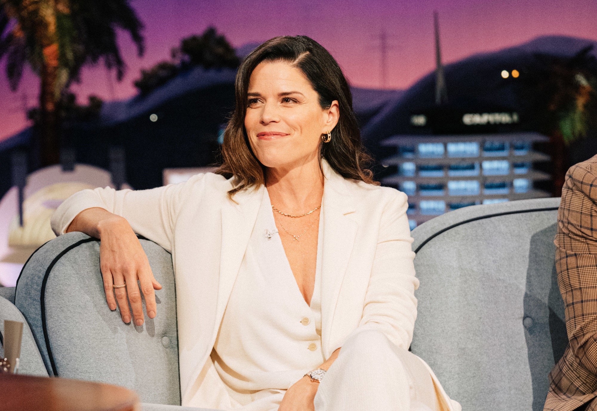 Neve Campbell in a suit on a couch