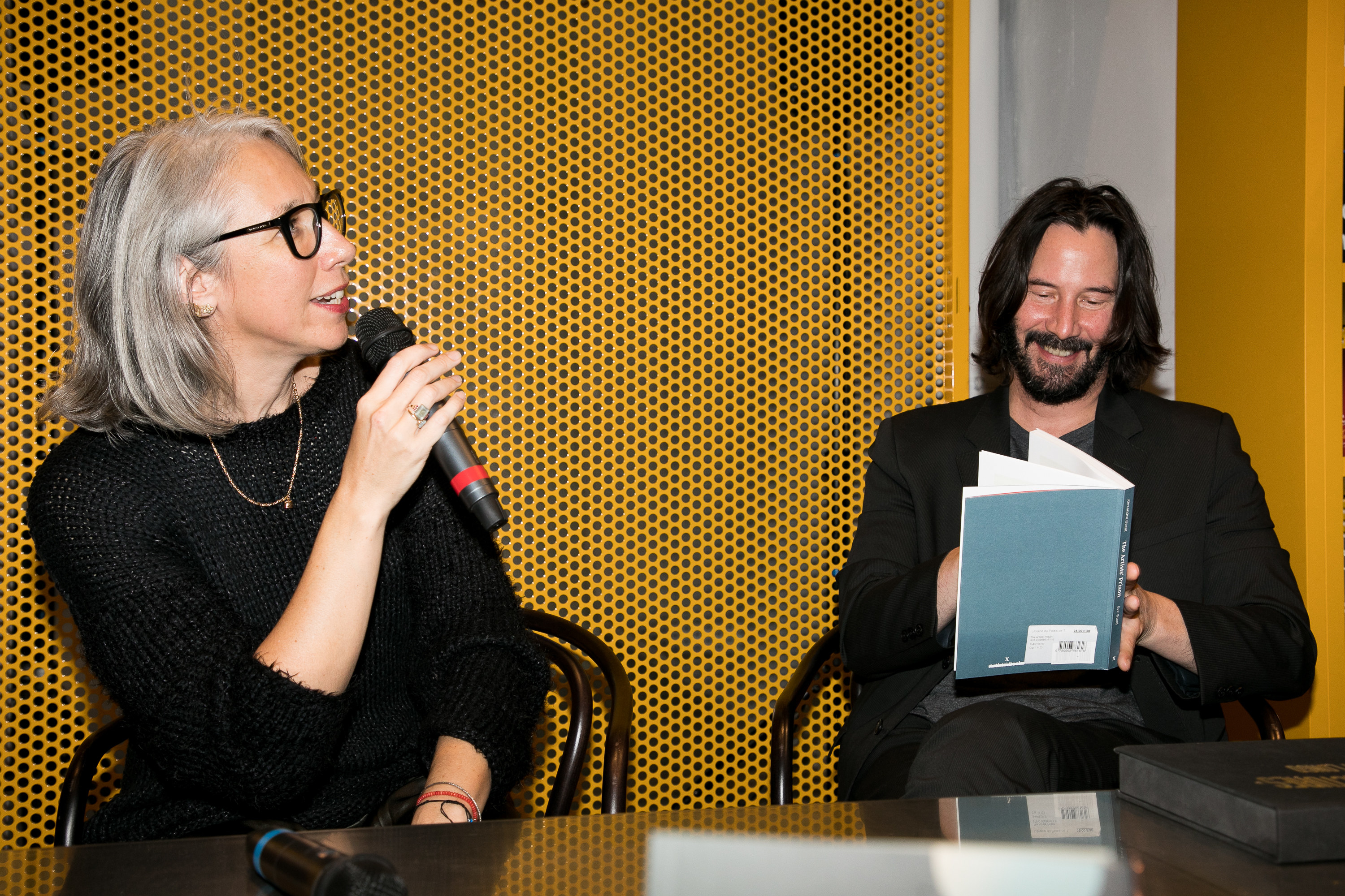 Alexandra speaks into a microphone while Keanu opens a copy of one of their books
