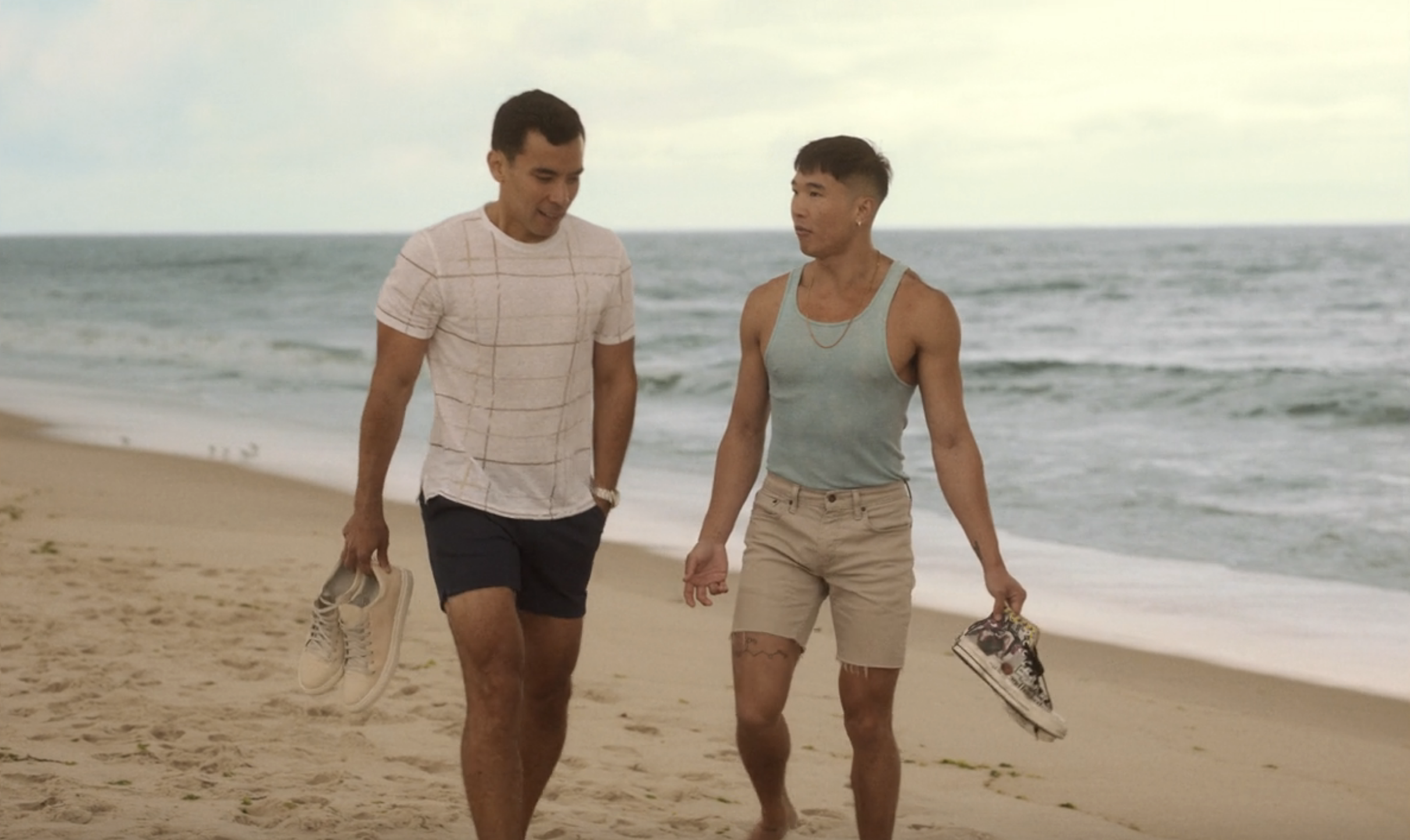 Ricamora and Booster walk along the sand on a beach holding their sneakers