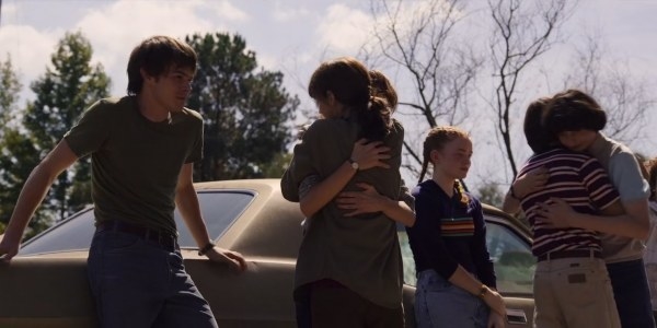 Girls and boys hug each other while some stand against a car