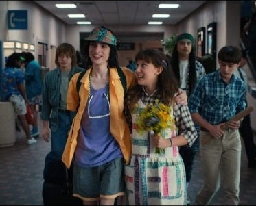 Kids walk in a school corridor. A boy has his arms around a girl who holds a bouquet of flowers