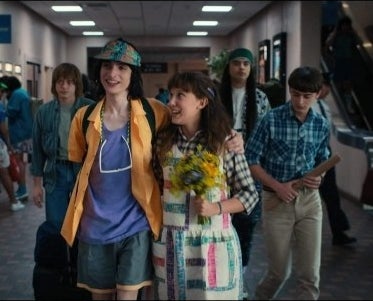 Kids walk in a school corridor. A boy has his arms around a girl who holds a bouquet of flowers