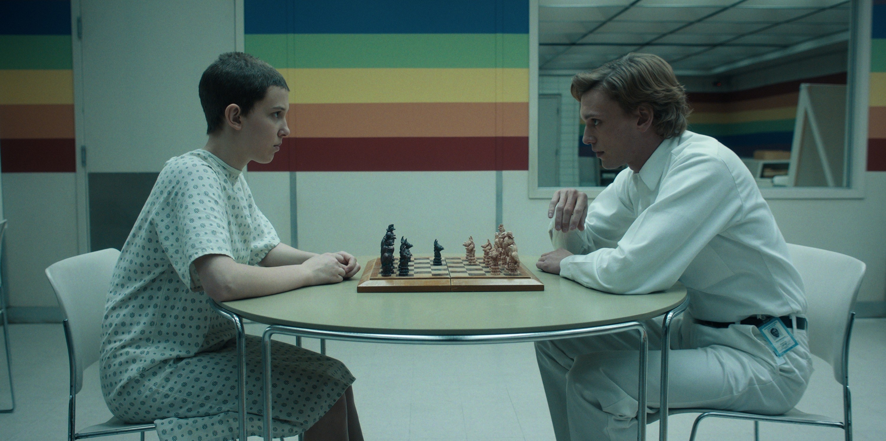 Eleven with her head shaved again wearing a hospital gown and playing chess with One who is wearing an administrators uniform