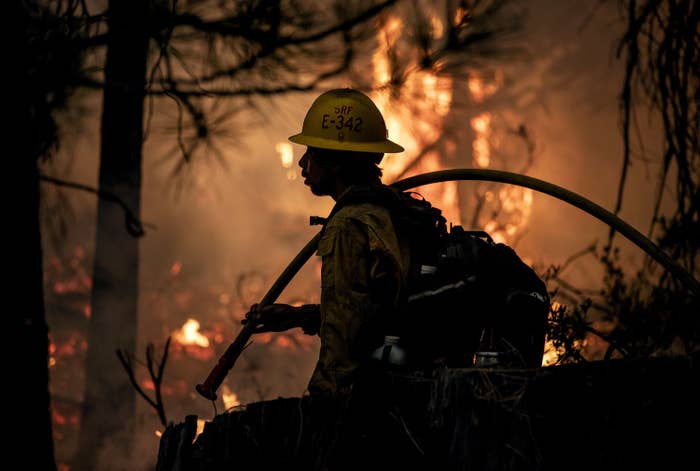 A firefighter wearing a helmet and carrying a hose is almost silhouetted by a blaze behind them