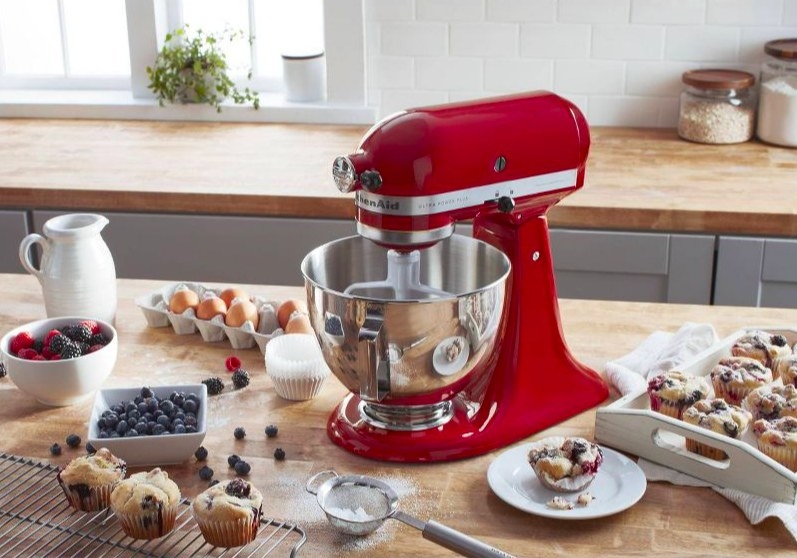 Red KitchenAid mixer on a kitchen counter next to baked blueberry muffins and various baking ingredients