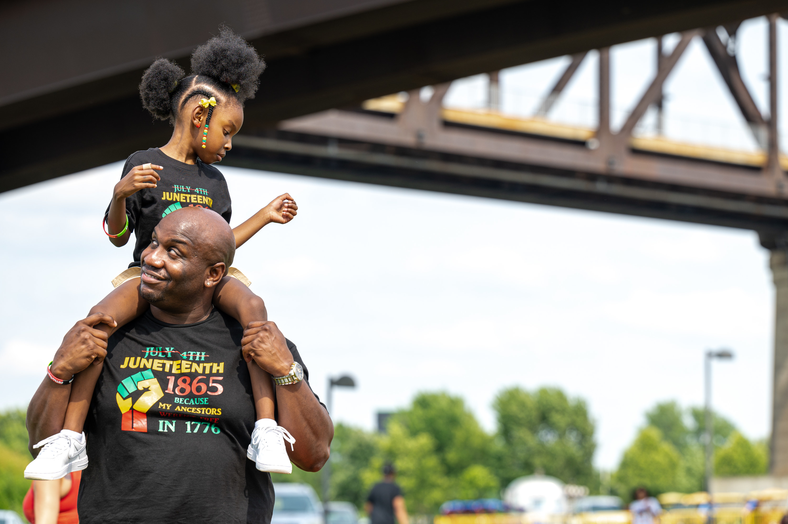 a father holding his daughter on his shoulders wearing Juneteenth shirts