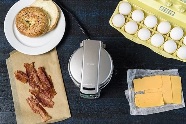 The sandwich maker with egg, bacon, and other ingredients next to t