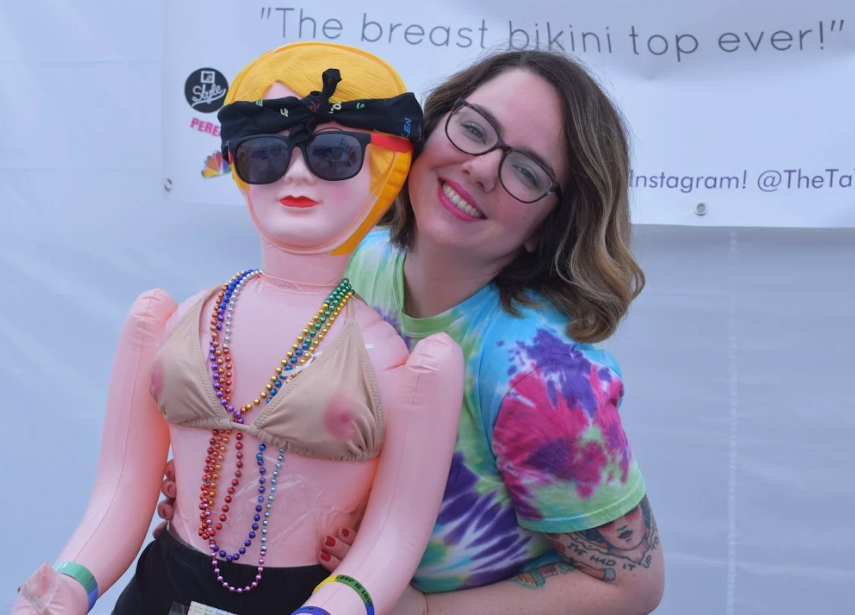 A smiling woman holds a blow-up doll wearing sunglasses and a bikini designed to look like breasts with nipples