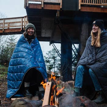 two models wrapped in rumpl blankets and sitting at an outdoor fire