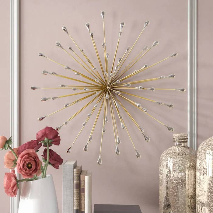 Golden starburst wall art with crystal tips mounted on pale pink wall with decorative books, jars, and flowers beneath it