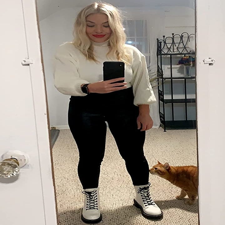 reviewer wearing the white boots with black pants and a white sweater while standing next to their cat