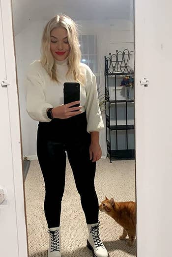 reviewer wearing the white boots with black pants and a white sweater while standing next to their cat