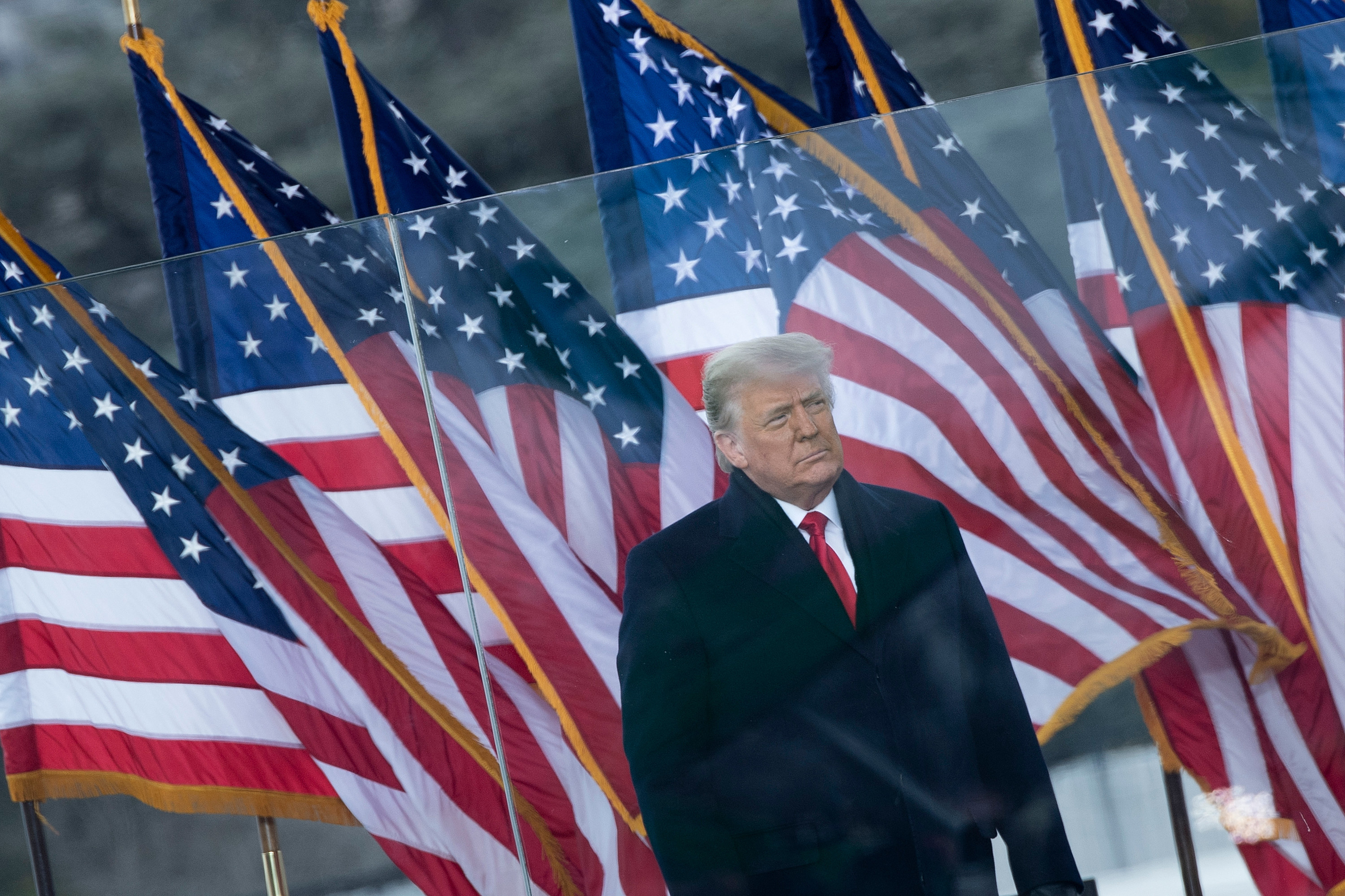 Trump wears a big jacket, red tie and stands in front of a row of six US flags behind a transparent partition