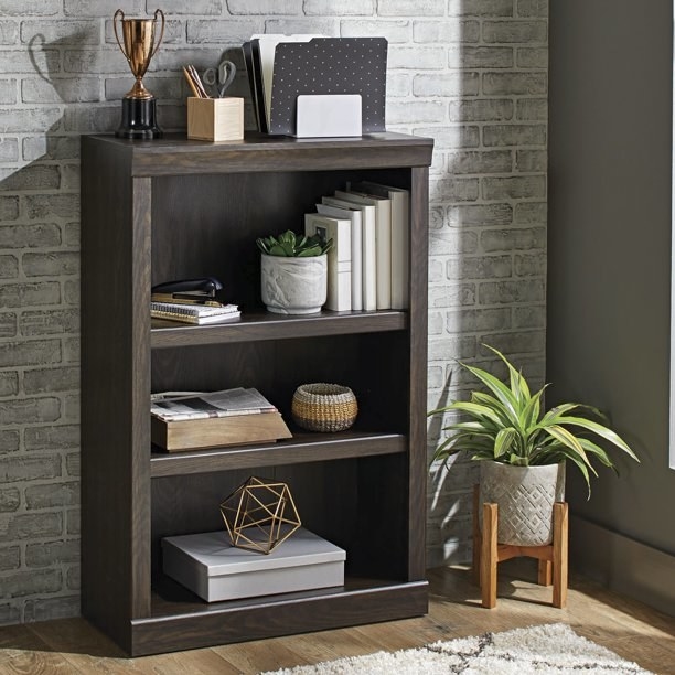 a three shelf bookcase in dark oat storing books and a houseplant