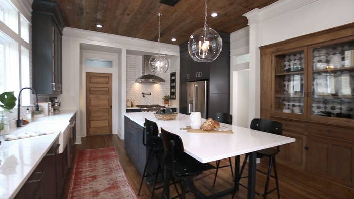 A kitchen designed by Joanna Gaines, including glass globe light pendants, a wood and glass china cabinet, and dark grey cabinets with a wood plank ceiling