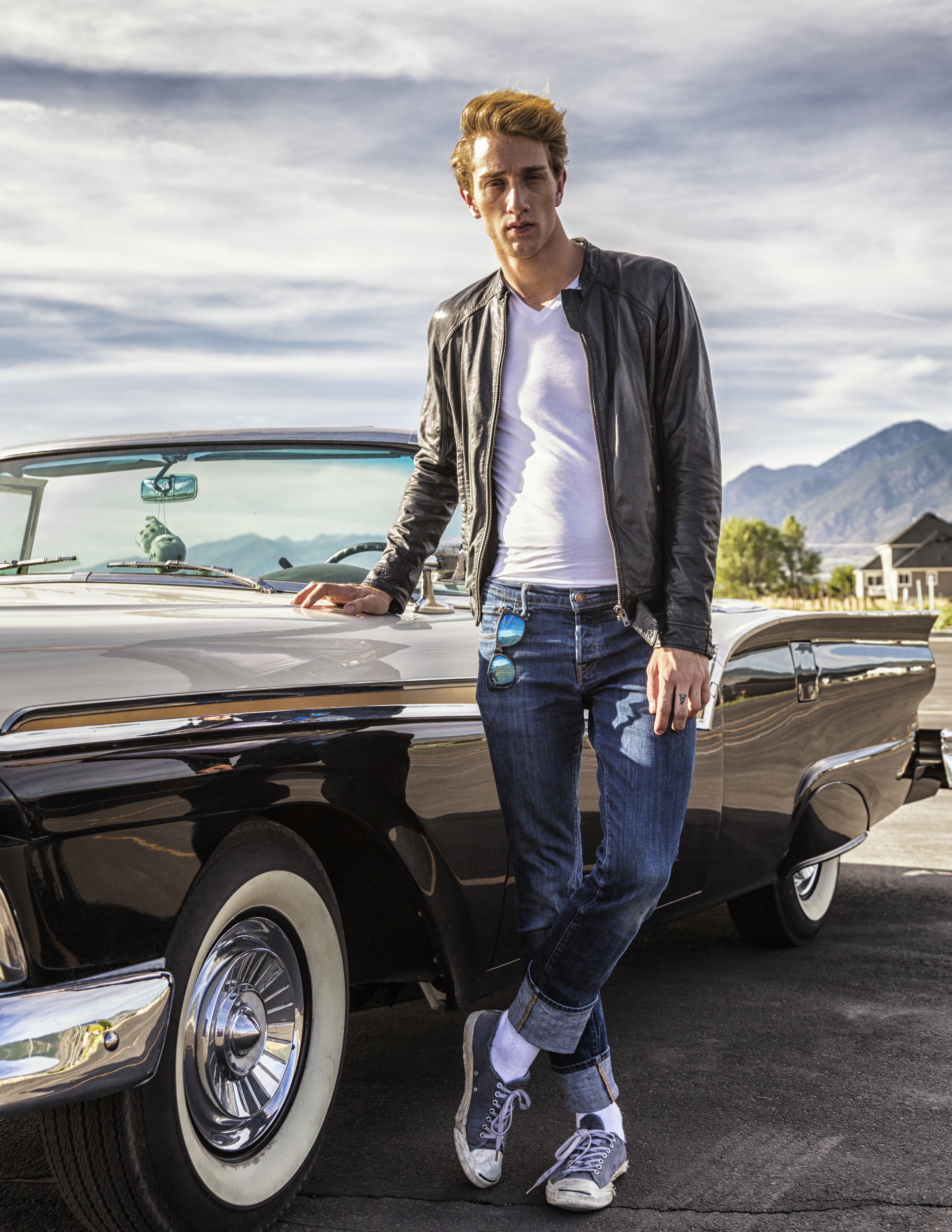 A man in a leather jacket poses next to a convertible