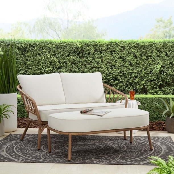 Wicker loveseat and ottoman with cream cushions out on a patio in front of greenery
