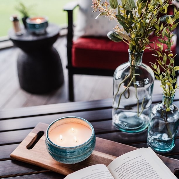 Lit citronella candle on a table with a book, a wooden board and vases with greenery