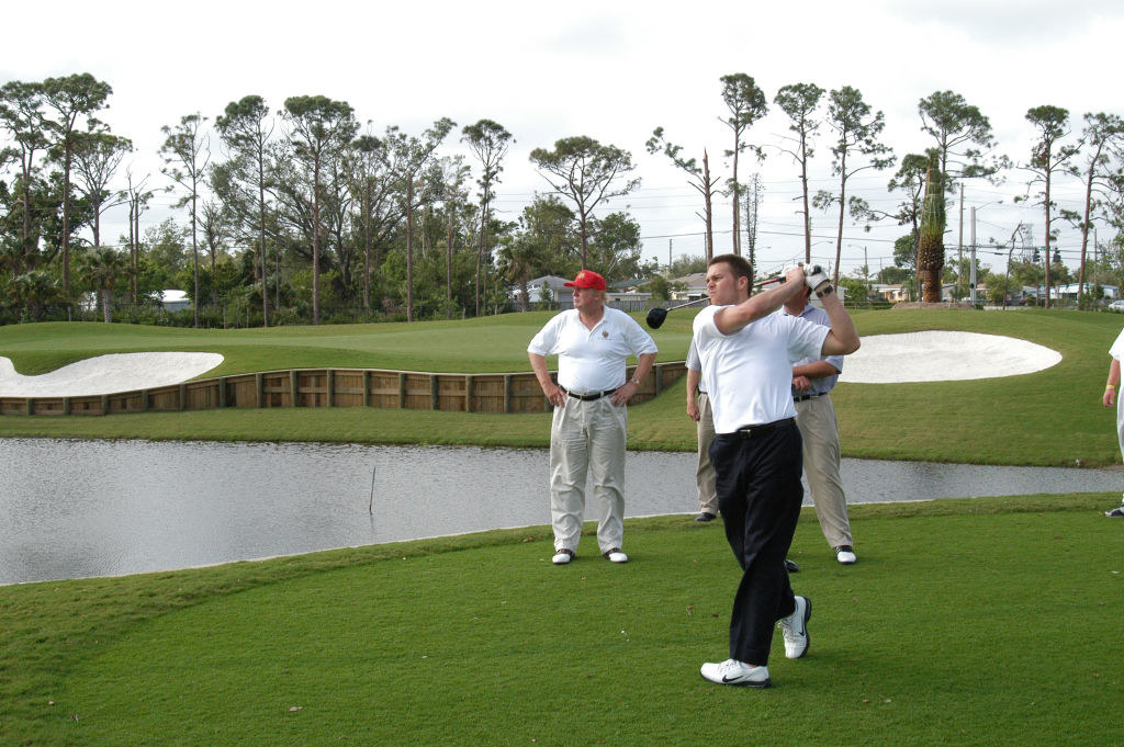 tom hitting a golf ball while trump stands to the side