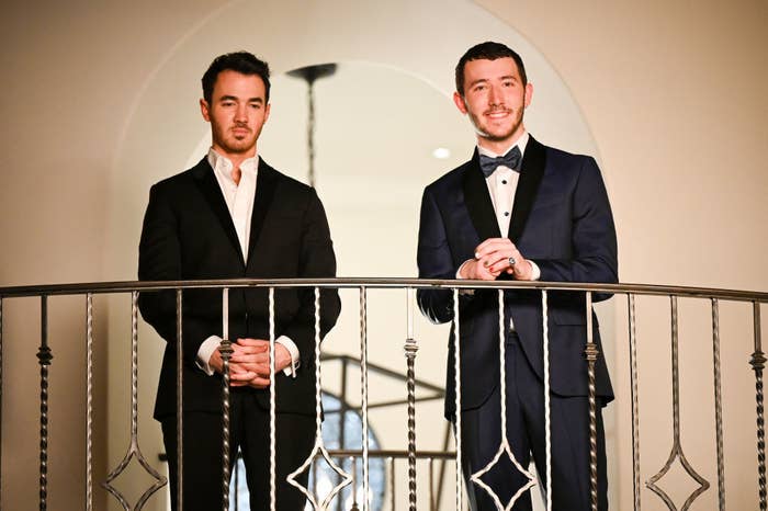 Kevin Jonas and Frankie Jonas standing on a balcony wearing suits
