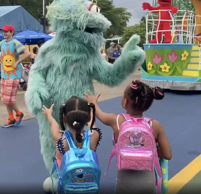Two little girls outstretch their arms while a large blue furry character points at someone in the crowd