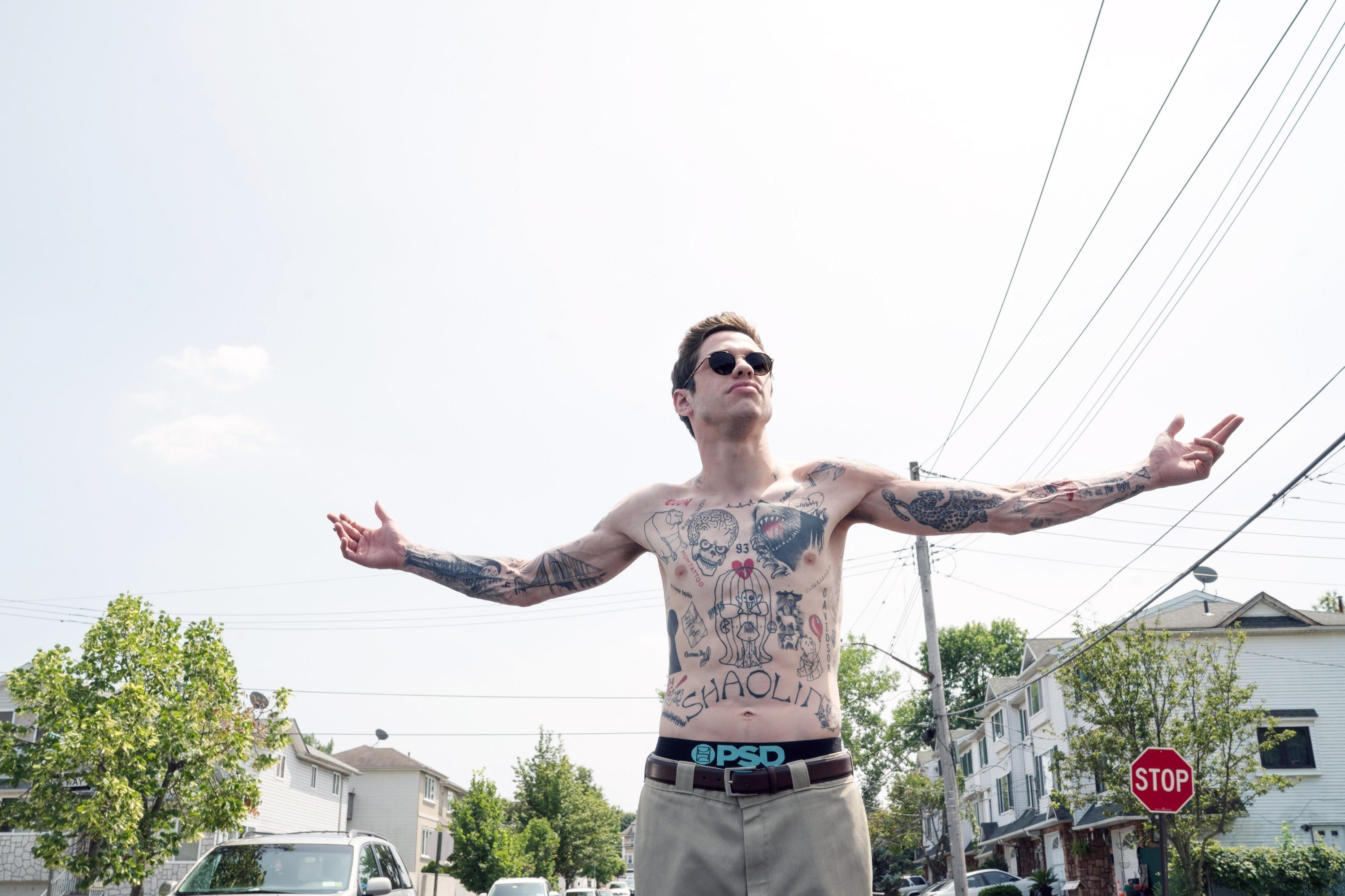 A shirtless Pete with many tattoos standing outside with his arms outstretched