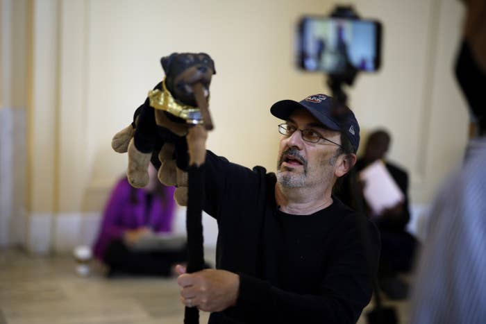 A man wearing glasses and a ball cap holds up a dog puppet with a cigar in its mouth