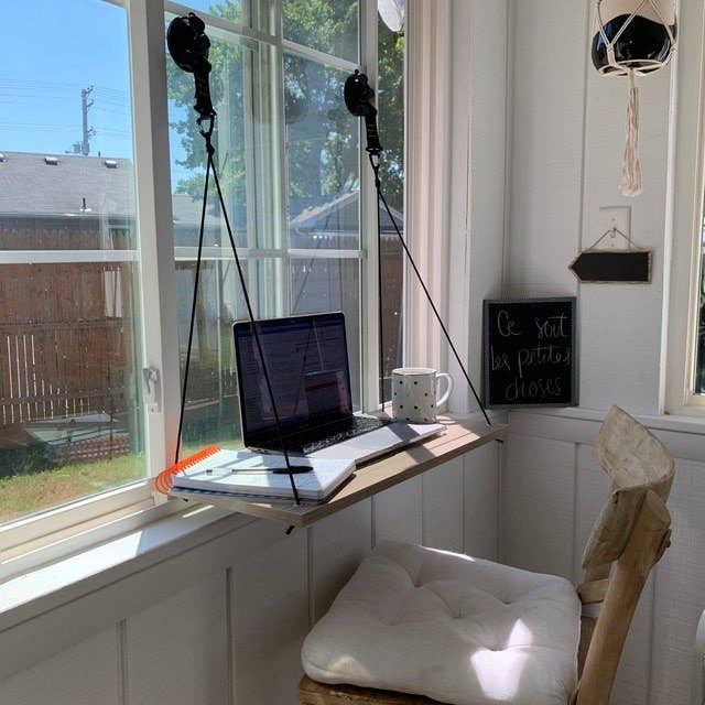 A wooden desk hung up by ropes and suction to a window with a laptop on it