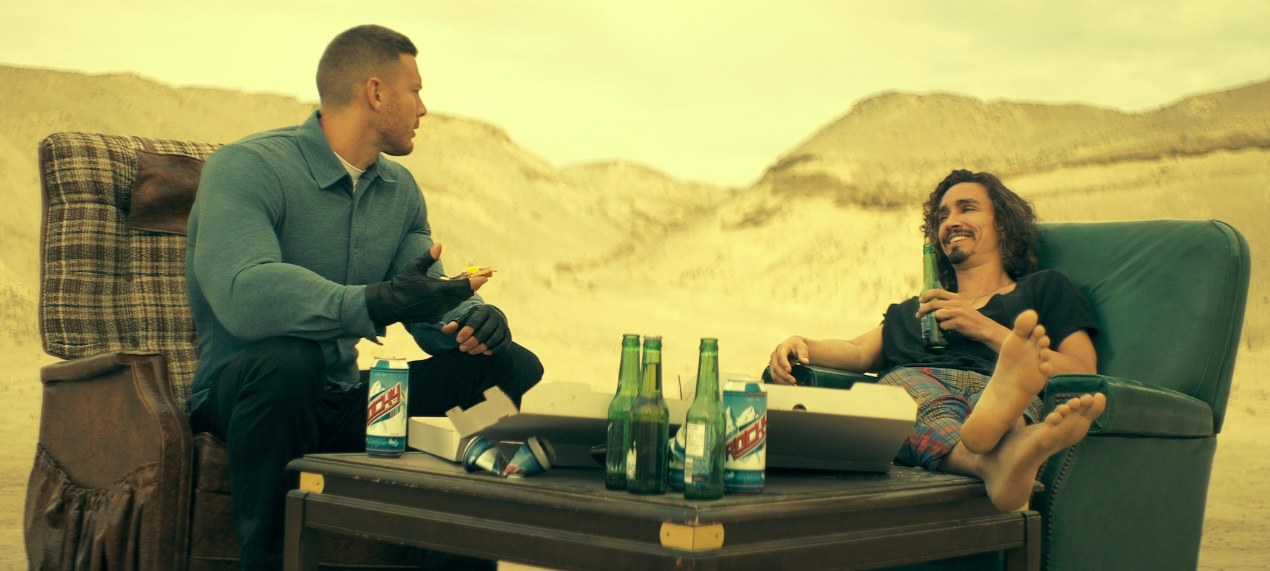 Luther and Klaus sit on couches eating pizza and drinking bear while talking in the middle of a desert