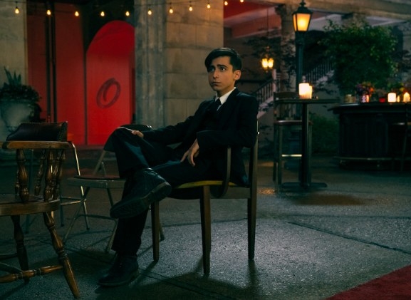 Five wears a suit and sits on a chair in a courtyard with a bar and party lights in the background