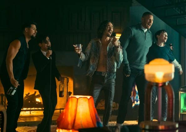 Diego, Five, Klaus, Luther and Viktor sing into microphones while holding alcohol and standing in a row in front of a fireplace in a hotel
