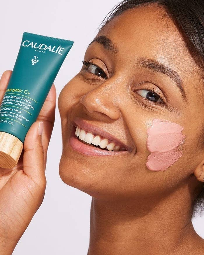 A person smiling while holding a tube of the mask with some of the product on their face