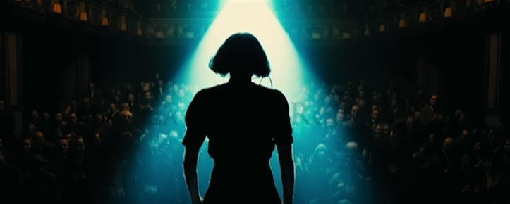 The silhouette of a woman onstage, taken from behind her so we see the spotlight flooding towards her and the packed audience in front of her. She has short hair in a voluminous bob